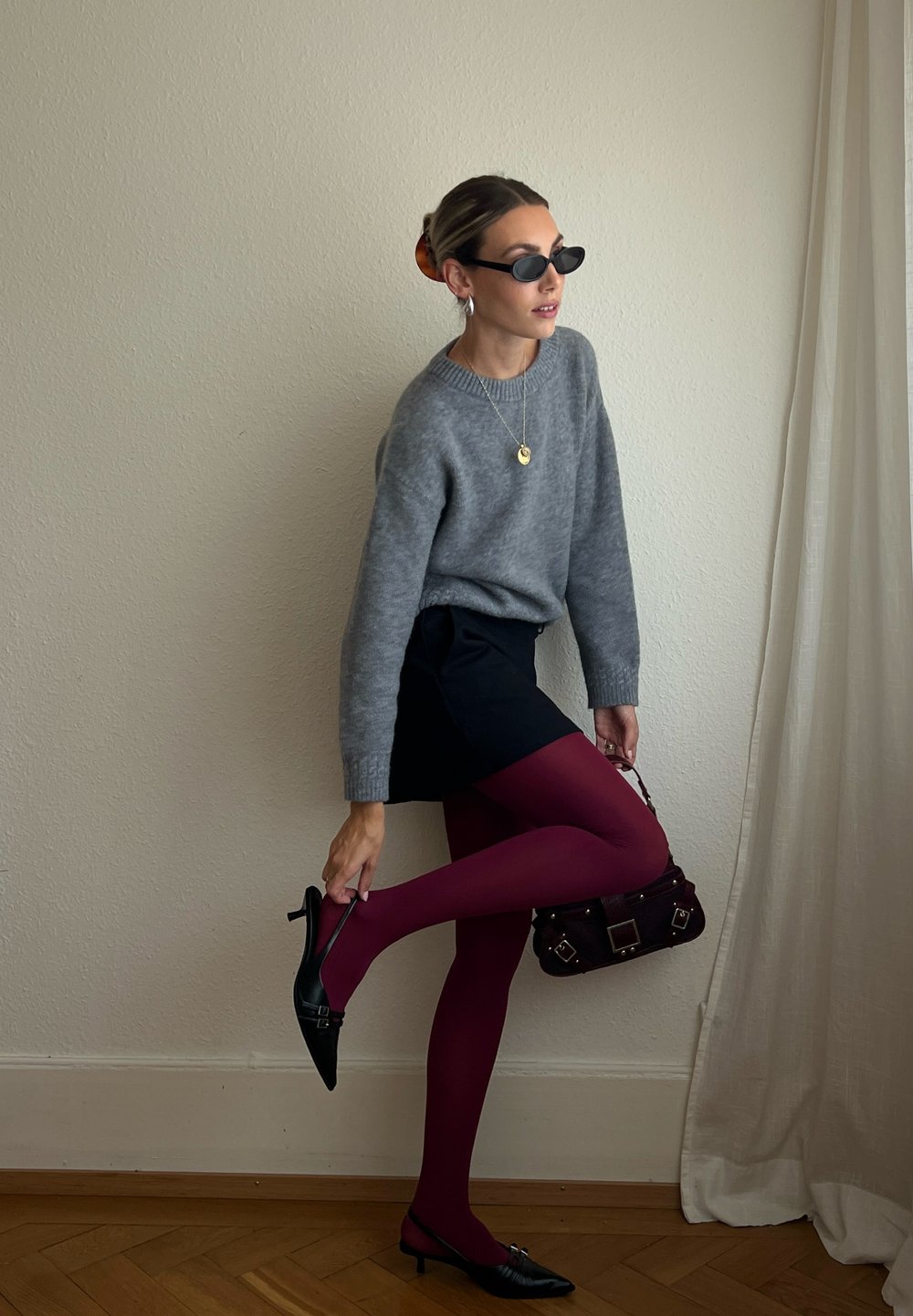 Pull gris, mini-jupe noire, collants bordeaux, chaussures à talons noires, petit sac à main foncé. Cadre intérieur avec des murs et un sol de couleur claire.