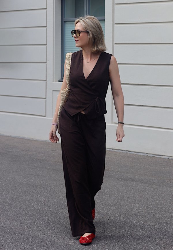 Žena s blond vlasmi, oblečená v čiernom top bez rukávov a červených topánkach, kráča po ulici pri bielej budove s oknom.
