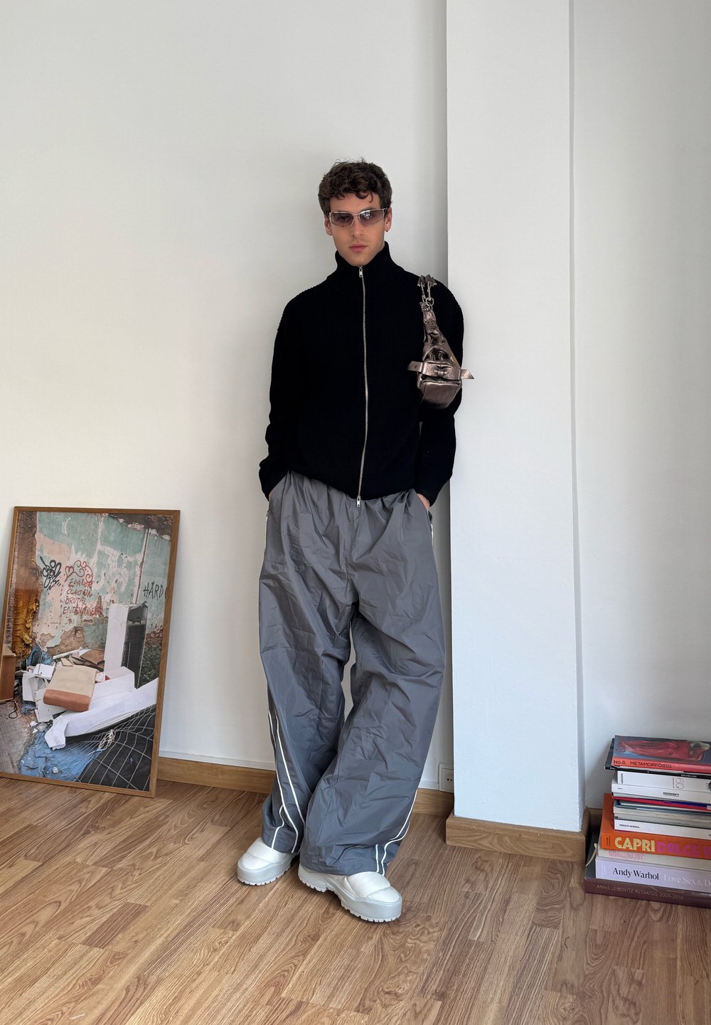Svart glidelås strikkegenser, grå brede bukser med sidestriper, hvite chunky sneakers og metallisk veske. Bakgrunnen har et foto og stablede bøker.