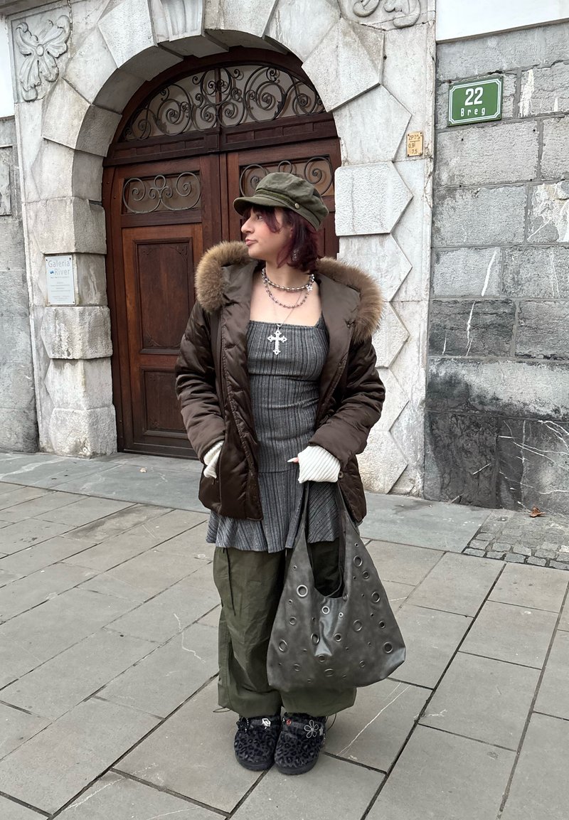 Veste matelassée marron avec bordure en fourrure, robe grise rayée, pantalon ample vert, chaussures noires duveteuses et sac cabas gris avec de grands œillets.