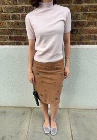 Lichtroze top met korte mouwen, tan suède rok met metallic bloemenaccenten, gecombineerd met grijze flats en een kleine zwarte handtas.