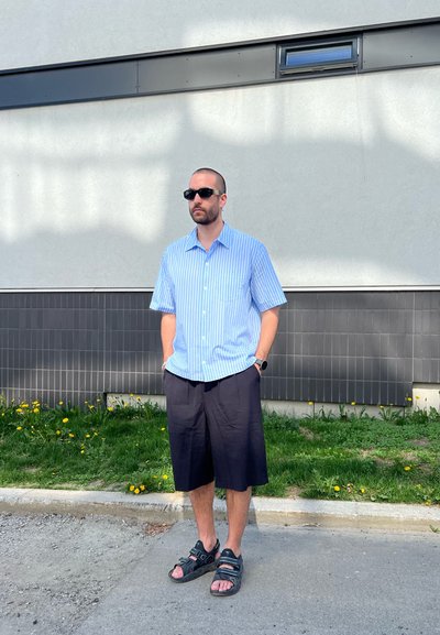 Camisa de botones de manga corta a rayas azul claro, pantalones cortos negros hasta la rodilla y sandalias negras con tiras. De pie contra una pared gris texturizada.