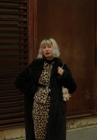Svart pelsoverlekk over en leopardmønstret kjole med belte, som holder en liten leke. Klippet, pastellfarget hår. Urban bakgrunn med treplanker.
