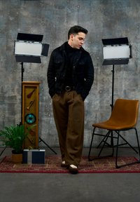 Homme en veste noire et pantalon marron se tient sur un tapis rouge entre une chaise marron et une plante, avec deux éclairages de studio et un affichage musical derrière lui.