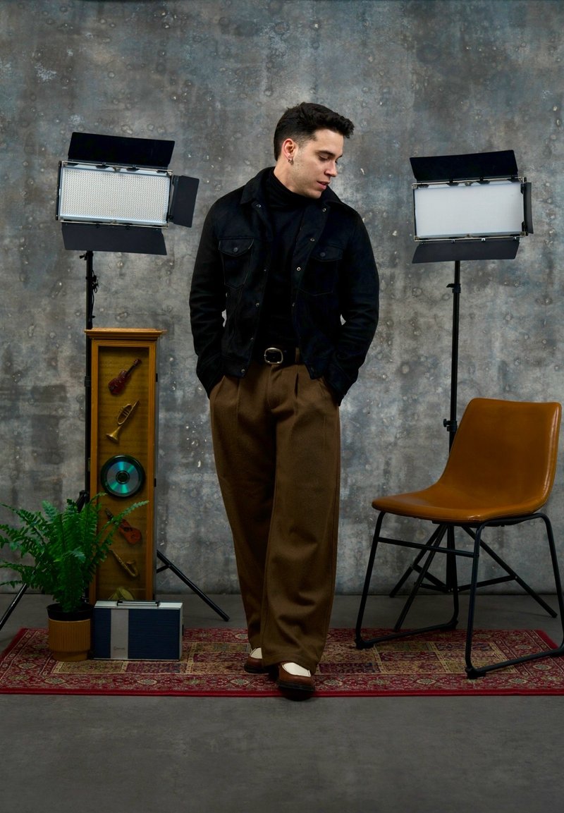 Mann in schwarzer Jacke und brauner Hose steht auf einem roten Teppich zwischen einem braunen Stuhl und einer Pflanze, mit zwei Studiobeleuchtungen und einer Musikanzeige hinter ihm.