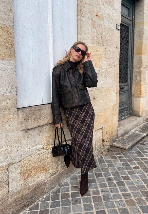 Una mujer con gafas de sol, chaqueta de cuero negra y falda de cuadros se ajusta las gafas mientras sujeta un bolso junto a un edificio de piedra.