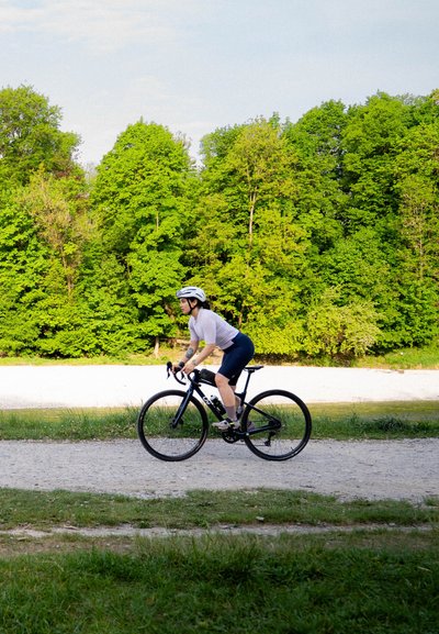 Bicicleta de carretera negra con neumáticos delgados, cuadro ligero y manillares en posición de caída. El ciclista lleva una camiseta blanca, pantalones cortos negros y un casco blanco.