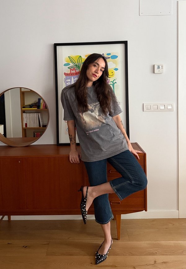 Femme aux longs cheveux foncés appuyée sur un buffet en bois, portant un t-shirt graphique gris, un jean court et des chaussures noires pointues avec des clous.