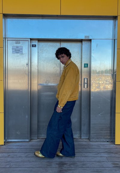 Sudadera amarilla con textura y cuello, combinada con jeans de pierna ancha de color azul oscuro. El modelo está de pie cerca de un ascensor de acero inoxidable.
