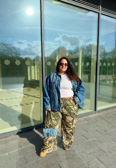 Chaqueta de mezclilla, top corto blanco, pantalones de carga camuflados y botas marrones. Bolso acolchado azul. De pie junto a una estructura de vidrio reflectante, con un fondo iluminado por el sol.