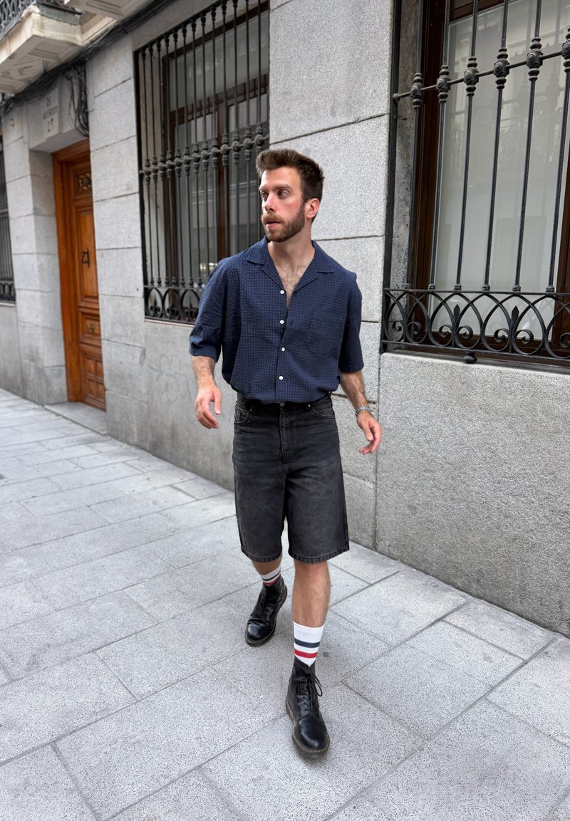 Homme marchant sur un trottoir en ville portant une chemise bleu marine à motifs, un short noir, des chaussettes rayées et des bottes noires près d’un bâtiment avec des fenêtres grillagées.