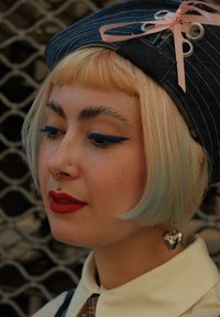 Korts denimkeps med rosa banddetalj, kort blont hår med blå accenter, djärv blå eyeliner, röda läppar och ett hjärtformat öra.