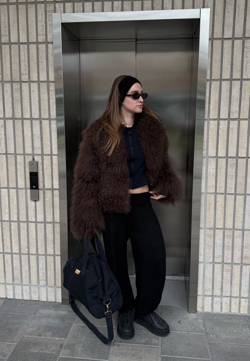 Veste en fausse fourrure marron, top noir court, pantalon noir à jambes larges, chaussures noires et sac noir. Debout près d'un ascenseur argenté aux murs texturés.