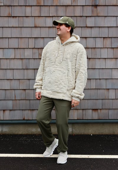 Hombre con sudadera con capucha de forro polar beige con estampado, pantalones verde oliva, zapatillas blancas y gorra verde, sonriendo frente a una pared de tejas marrones.