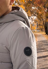 Ανοιχτό γκρι φουσκωτό μπουφάν με κουκούλα, που διαθέτει ένα στρογγυλό μαύρο λογότυπο στην αριστερή μανσέτα. Λείος υφασμάτινος τύπος, κομψός σχεδιασμός.