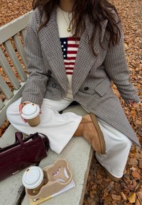 Manteau croisé gris à carreaux porté sur un pull crème avec un motif du drapeau américain, pantalon blanc, bottes UGG marron clair et une tasse de café.