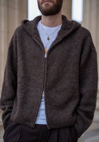 Sweat à capuche marron zippé, fabriqué en matière texturée. Comprend une capuche, une fermeture éclair à l'avant et des poignets côtelés, superposé sur une chemise bleu clair.