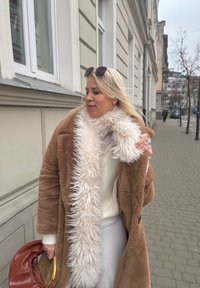 Ξανθιά γυναίκα με καφέ γούνινο παλτό και αφράτο άσπρο κασκόλ προσαρμόζει το γιακά της ενώ περπατά σε πεζοδρόμιο της πόλης κοντά σε μπεζ κτίρια.