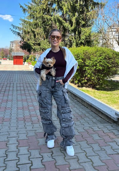 Pantalones cargo en denim gris con múltiples bolsillos, combinados con una blusa oscura y un chal blanco. Un perro pequeño con arnés es sostenido en los brazos.