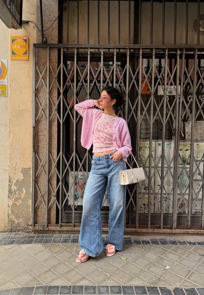 Mujer con cárdigan rosa, top floral, vaqueros anchos, sandalias rosas y gafas de sol posa junto a una puerta metálica cerrada, sosteniendo un bolso blanco en la acera de la ciudad.