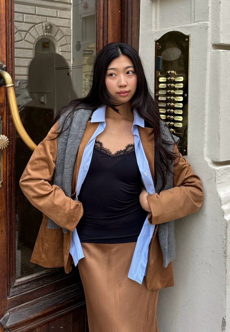 Giovane donna con lunghi capelli neri, indossa una giacca marrone a strati e una camicia blu, si trova accanto a una porta di legno con pulsanti per citofono.