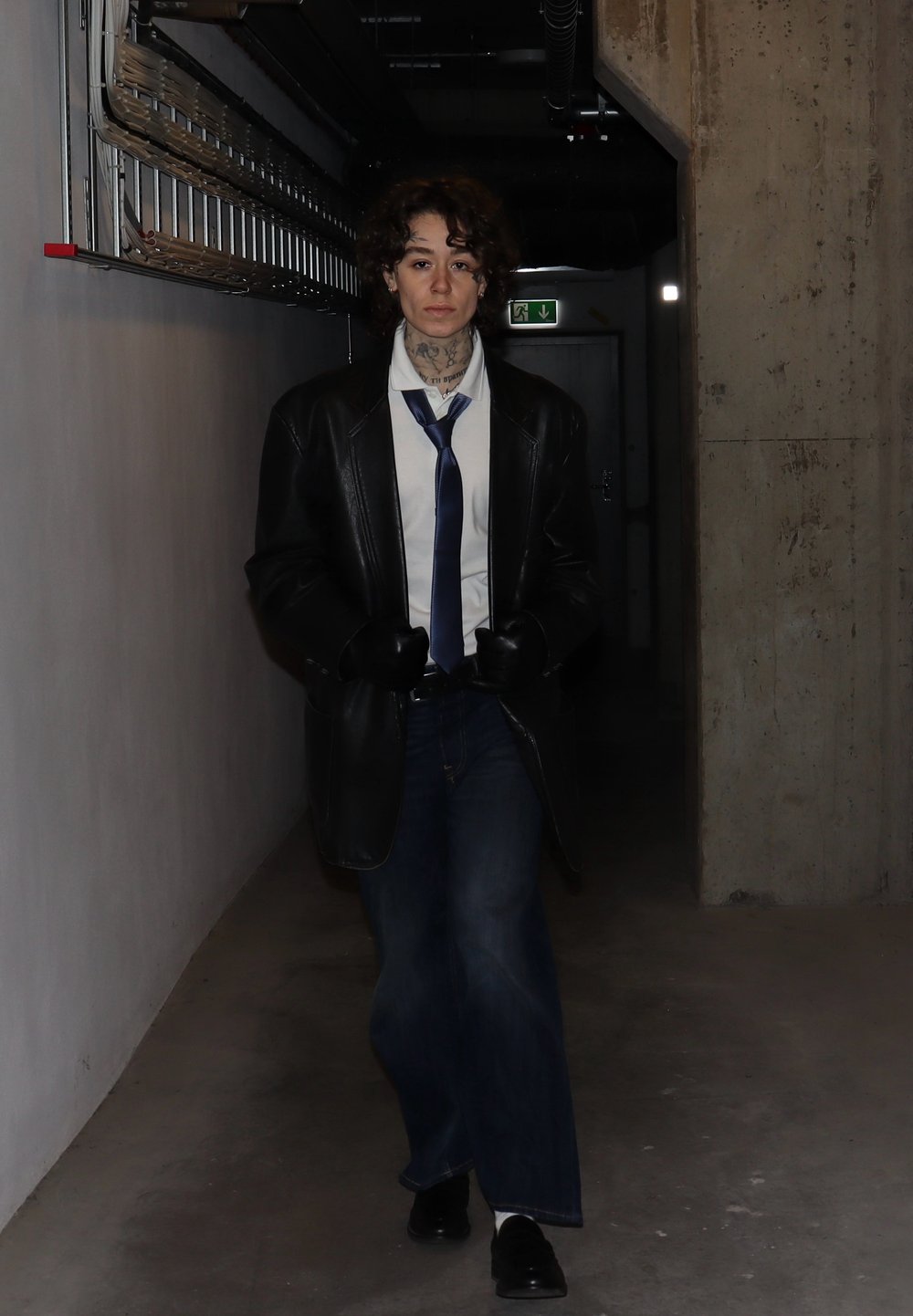 Czarny skórzany płaszcz na białej koszuli, niebieskim krawacie i ciemnych dżinsach. Ciemne rękawiczki i buty. Kręcone włosy, tatuaż na szyi, stojący w korytarzu.