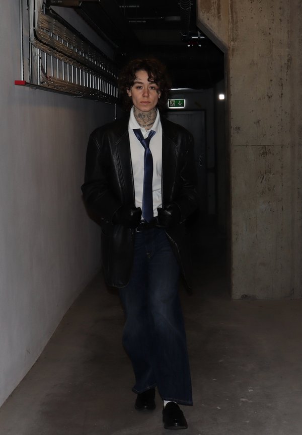 Manteau en cuir noir sur une chemise blanche, cravate bleue et jean foncé. Gants et chaussures noirs. Cheveux bouclés, cou tatoué, debout dans un couloir.