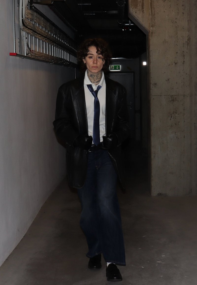 Zwarte leren jas over een wit hemd, blauwe das en donkere jeans. Donkere handschoenen en schoenen. Gekruld haar, getatoeëerde nek, staand in een gang.