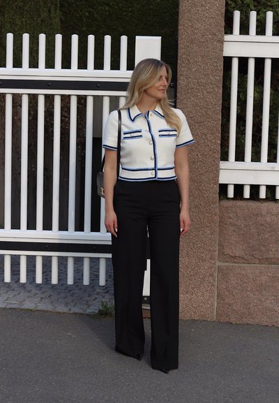 Chaqueta corta de tweed blanco con acentos azules, combinada con pantalones de pierna ancha negros. De pie cerca de un fondo de cerca blanca.