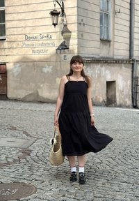 Abito nero a balze senza maniche, in tessuto testurizzato. Borsa in paglia intrecciata con dettaglio floreale, scarpe nere chunky, calzini bianchi con accenti rossi. Sfondo in ciottoli.