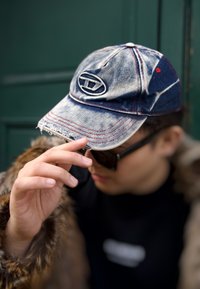 Džínová baseballová čepice s opraným okrajem, modrou látkou, červeným prošíváním a logem na přední straně. Částečně viditelné černé sluneční brýle a kožich.