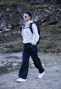 Vrouw met een zonnebril, een lichte jas, zwarte broek en sneakers wandelt over een rotsachtig pad met een zwarte rugzak.