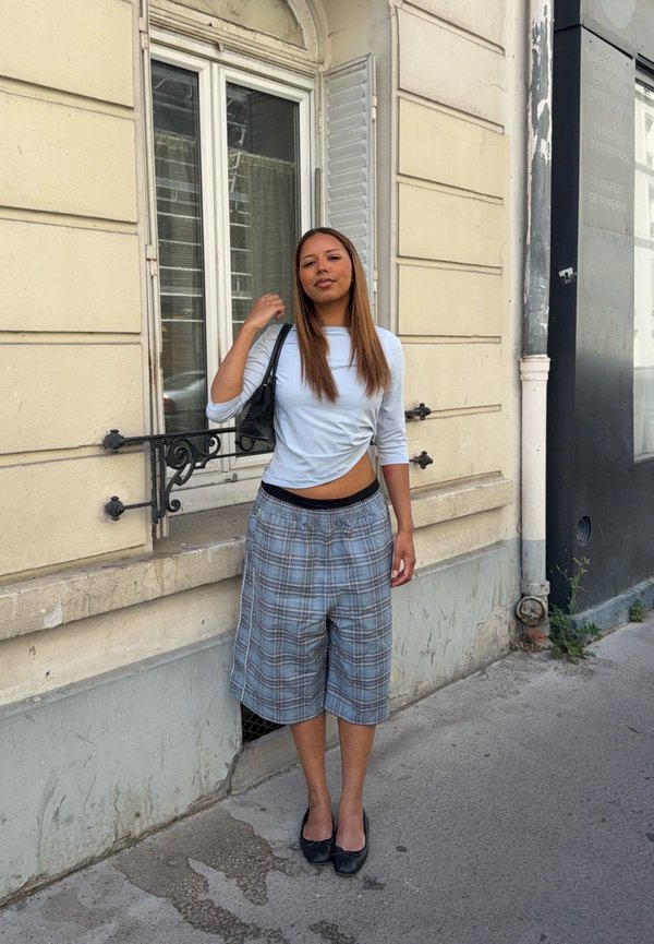 Femme aux longs cheveux, portant un haut blanc et un short large à carreaux, se tient sur le trottoir près de la fenêtre d’un bâtiment beige, tenant un sac à main noir.