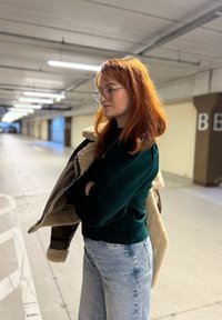 Une personne se tient dans un parking, portant un sweat vert, un jean large bleu clair, et tenant une veste marron doublée en peau retournée.