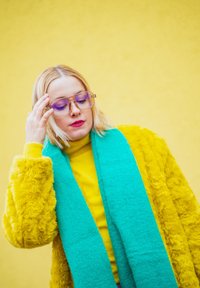 Umělé kožešinový žlutý kabát, zelený šál, žlutý svetr. Čiré rámečky brýlí, blond vlasy, stojící na žlutém pozadí.