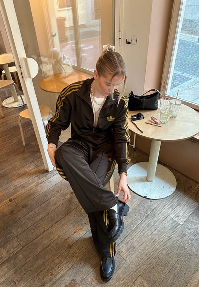 Chándal de color marrón oscuro con rayas amarillas, que incluye chaqueta con cremallera y pantalones con cintura elástica. Zapatillas negras con suelas gruesas. Suelo de madera natural.