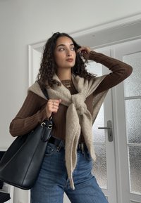 Brun ribstrikket langærmet trøje, beige tørklæde knyttet foran, denimjeans, sort lædertaske med gyldne detaljer, stående indendørs ved en dør.