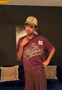 Νέος άνδρας που φορά κίτρινο καπέλο και μπλούζα μαρόν με την επιγραφή "Los Angeles" και αριθμό 93, στέκεται μέσα σε κλειστό χώρο μπροστά σε σκούρο τοίχο και μαξιλάρια με σχέδια.