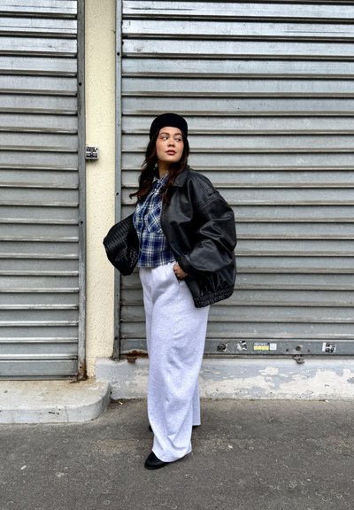 Mujer con boina negra, chaqueta de cuero negra, camisa de cuadros azul, pantalones anchos blancos, sosteniendo un bolso tejido negro, de pie junto a persianas metálicas cerradas.