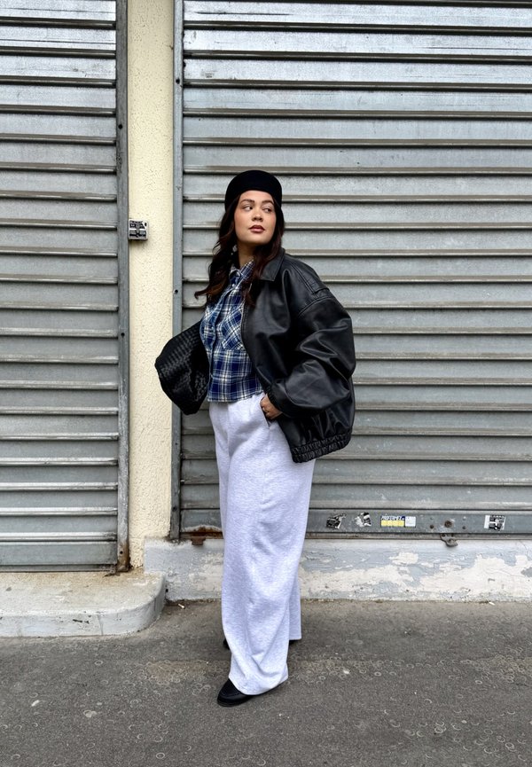 Kobieta w czarnym berecie, czarnej skórzanej kurtce, niebieskiej kratkowanej koszuli, białych szerokich spodniach, trzymająca czarną plecioną torbę, stojąca obok zamkniętych metalowych rolet.