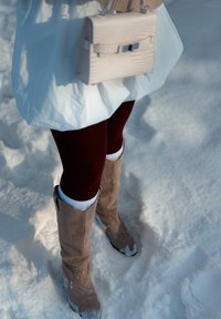 Bottes hautes jusqu'au genou en daim beige, leggings bordeaux, jambières blanches en tricot, et un petit sac à main clair sur un fond enneigé.