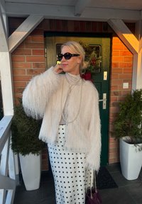 Femme blonde portant des lunettes de soleil, une veste blanche duveteuse, un collier de perles, une jupe à pois, tenant un sac à main bordeaux, debout près d'une porte verte.