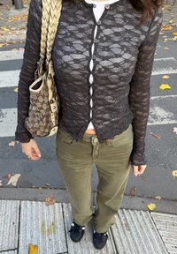 Crna čipkasta bluza s dugim rukavima i prednjim gumbima preko bijele košulje, maslinasto zelene manšestarske hlače i crne cipele s bijelim detaljima.