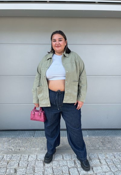 Camisa exterior verde oliva, top corto gris, pantalones oscuros de denim holgados, botas plataforma negras y un pequeño bolso rosa. Texturas suaves, ajuste relajado.