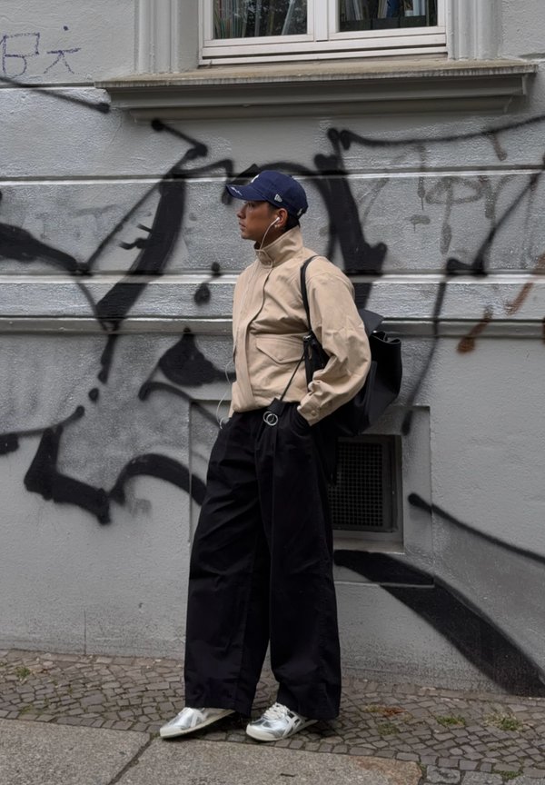 Osoba w beżowej kurtce i szerokich czarnych spodniach stoi przy ścianie pokrytej graffiti, z słuchawkami w uszach, z czarnym plecakiem i granatową czapką.