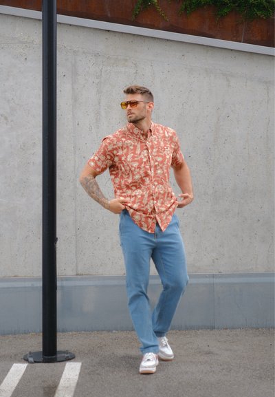 Camisa de manga corta en color coral con un patrón floral, combinada con jeans de color azul claro y zapatillas blancas. El modelo lleva gafas de sol y está de pie junto a un poste.