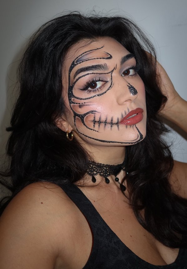Faccia dipinta con un design scheletrico nero, con accenti lucidi e glitter. Indossato con un choker nero e un top scuro, capelli lunghi e scuri visibili.