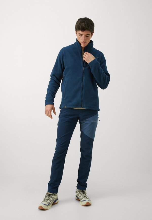 Navy Fleecejacke mit hohem Kragen, Frontreißverschluss und zwei Taschen; kombiniert mit dunkelblauen Hosen mit einem hellblauen Seitenstreifen und beigen Schuhen.