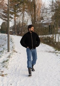 Homme portant une veste noire, un jean bleu et des bottes noires, marchant sur un chemin enneigé avec des arbres et des maisons en arrière-plan.