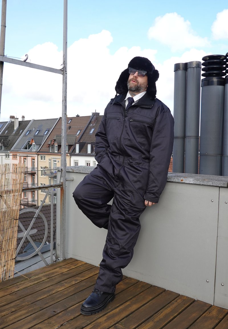 Hombre vestido con un overol aislante negro, gorro de piel, gafas de sol y camisa de vestir con corbata, apoyado en la barandilla de una azotea con edificios al fondo.